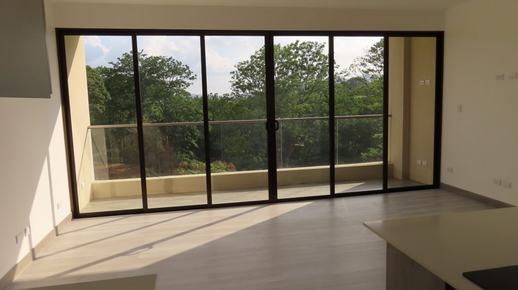 Las puertas corredizas de un apartamento nuevo. Las puertas corredizas son de vidrio y luego bordeado de metal diseñado para abrirse y cerrarse fácil. Se aprecia el bosque en la vista. El apartamento es de tonos blancos, beiges y grises.